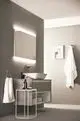 Badezimmeransicht mit hellgrauer Wand, umweltfreundlichem Waschbecken, Armatur und einem beleuchteten Spiegel. Ideal Standard Produkte sind integriert. Handtücher und Dekor sind vorhanden.
